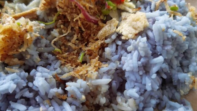 Close-up panning shot of Nasi Kerabu, a traditional Malaysian dish featuring butterfly pea flower blue rice served with shredded herbs, toasted coconut, chili paste, and salted egg.