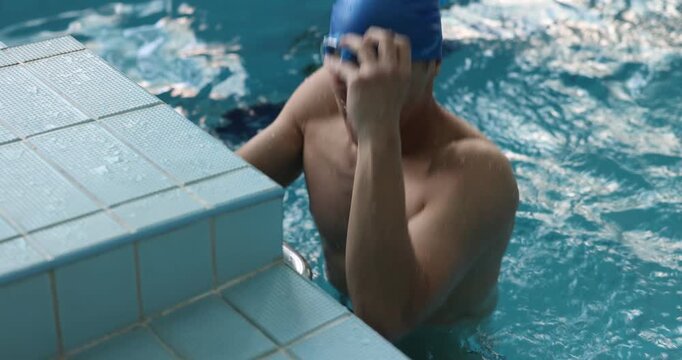 Young man in cap and goggles swimming near starting block in pool