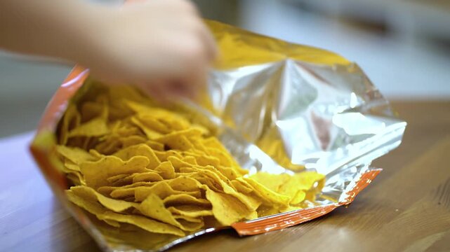 Close-up of female hands with red nails reaching into an open bag of golden nacho chips on a wooden table. Perfect stock footage for food, snacks, and lifestyle projects showing casual snacking and cr