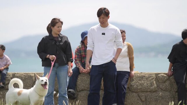 Young couple walks with dog near seaside wall. Group of people relaxes nearby, enjoying coastal view. White dog wags tail, joyful and playful. Peaceful seaside moment