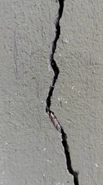 Close-up macro shot of a deep, long vertical crack running down a textured concrete wall with painted gray or olive surface.