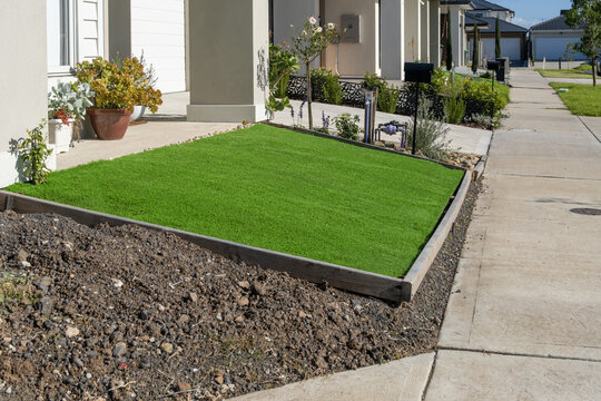 A patch of vivid synthetic turf or fake grass in a raised garden bed with timber edging beside suburban footpath. A modest and low-maintenance landscaping typical in new-build Australian front yards.