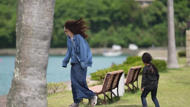 Woman walks briskly past palm trees near water. Child follows closely behind on grassy path. Bench sits idle near tranquil lake. Scene suggests a calm, natural park setting