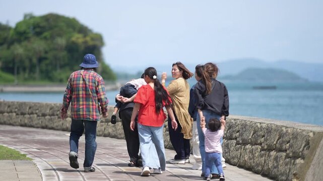 People stroll along seaside path. Man with a camera walks beside others. They pause to observe the water and distant hills. Sunlight glows softly over the calm sea. Perfect for travel, family