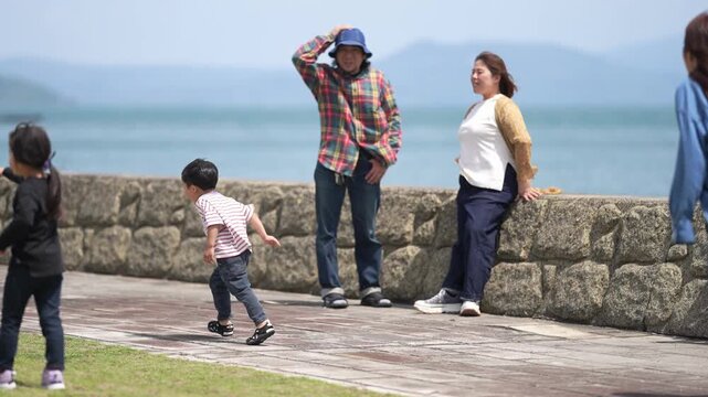 People stroll near seaside stone wall. Man shields eyes from sun, child runs nearby. Woman smiles, enjoying coastal walk. Scene captures casual seaside leisure. Perfect for travel, family, vacation