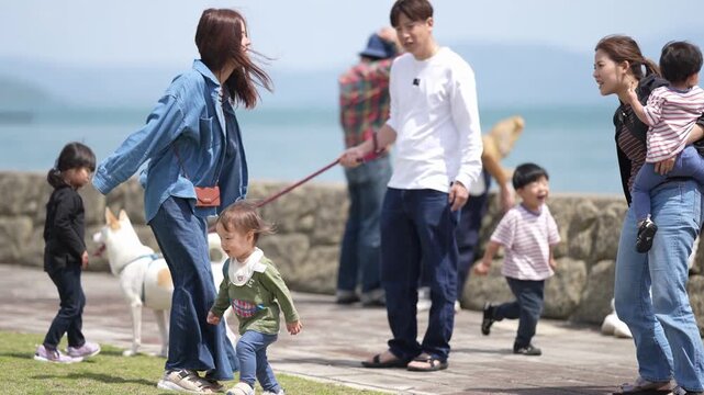 Woman laughs while walking with child and dog. Child runs beside her, chasing the dog. Group strolls along seaside path together. Sunlit day, relaxed, joyful atmosphere. Perfect for family, leisure