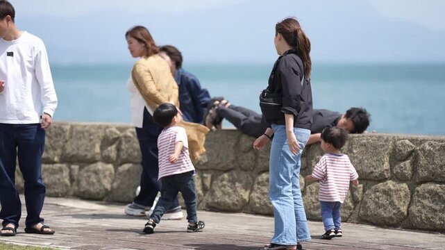 Coastside family strolls near stone wall. Parents carry children, one leans on stone. One adult rests against the wall. Children run ahead, curious and carefree. Lush blue sea stretches behind them