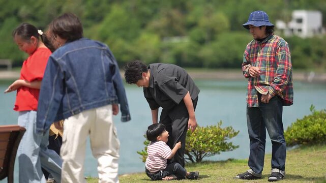 Child sits on grass near water. Adults gather, showing concern and care. Scene suggests a calm, outdoor moment of attention. People's focus points to the child's need for help