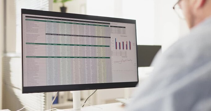 Accountant manager man in office computer spreadsheet analysis. Focused worker reviews finance reports and budget charts on a wide monitor. Tracking workload. Clear concept of financial stress.