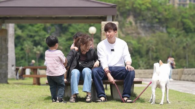 Family relaxes with dogs in a park. Mother, father, and child sit together. Child reaches for mother's face. Dog stands nearby, looking at camera. Scene captures gentle, peaceful moments