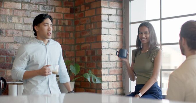 Diverse coworkers holding coffee mugs and laughing in loft break area, left man telling joke