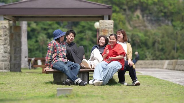 Four friends laugh together on a park bench. One wears a blue hat, another in red. They sit close, sharing smiles and stories. Green grass and trees frame their relaxed moment