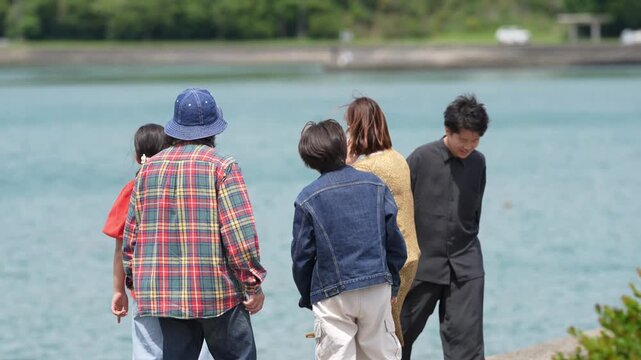 Group strolls along calm shoreline, enjoying quiet waterside. Soft light bathes them as they walk together, smiling, chatting. Lush green hills frame peaceful coastal scene behind them