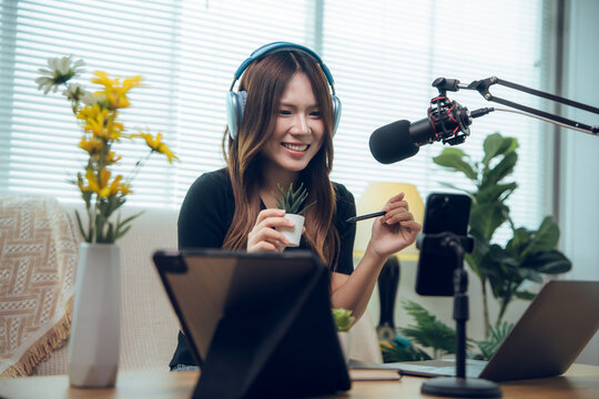 Cheerful Asian female content creator recording video blog on smartphone, holding small plant, live streaming from home studio setup with laptop, tablet, and microphone for social media channel.