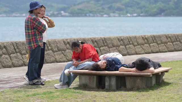 Three people relax near a calm sea. One stands, observing others. Two lie on a wooden bench. One person lies on their back. Another leans forward, appearing tired. Third rests face down