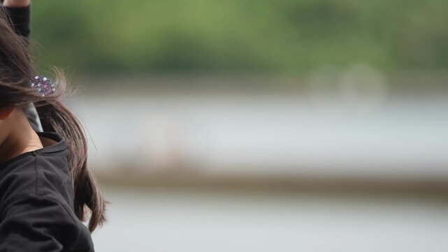 Small child gazes thoughtfully at water's edge. Dark hair flows gently in breeze. Black shirt contrasts soft, blurry background. Child's quiet contemplation evokes calm solitude