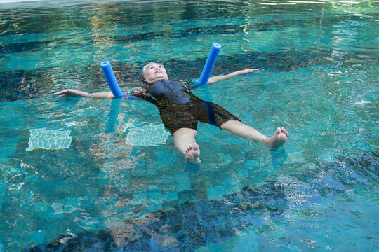 Senior woman floating in a swimming pool with pool noodles for aquatic therapy. Active elderly female doing water exercises for rehabilitation and healthy lifestyle.