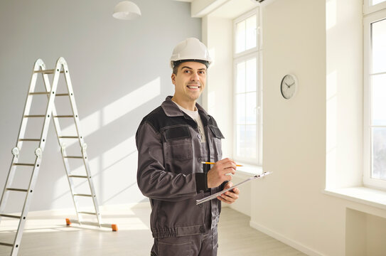 Contractor builder worker with helmet and clipboard. Handyman and engineer note repair and safety indoors, smiling in bright room near ladder. Clear reliable renovation and planning concept.