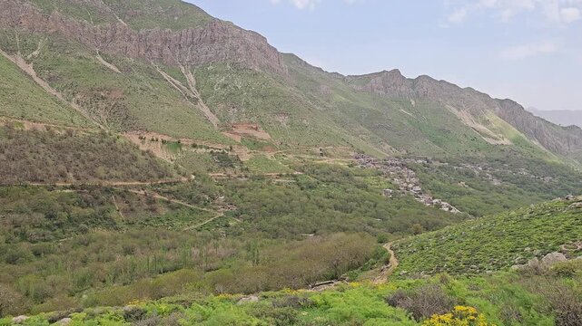 Spring wild herbs and yellow blossoms cover a green hillside leading toward a distant mountain village, framed by rugged alpine ridges under a partly cloudy sky.
📍 Kamalla village, Hawraman Takht, Ku