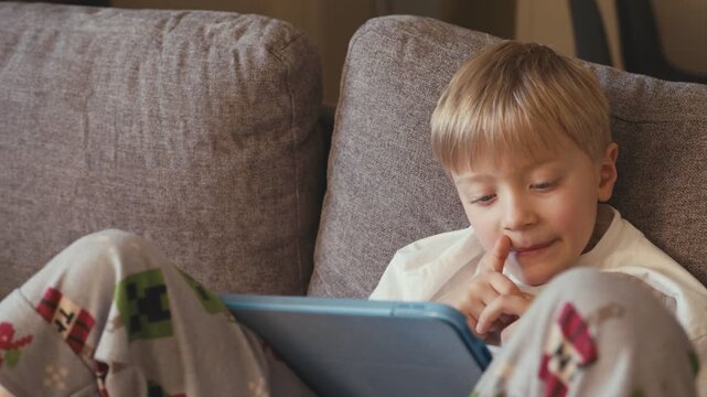 Little boy in pajamas playing with tablet on sofa