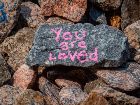 A grey stone painted with the pink message You are loved, resting among a pile of natural granite landscaping rocks