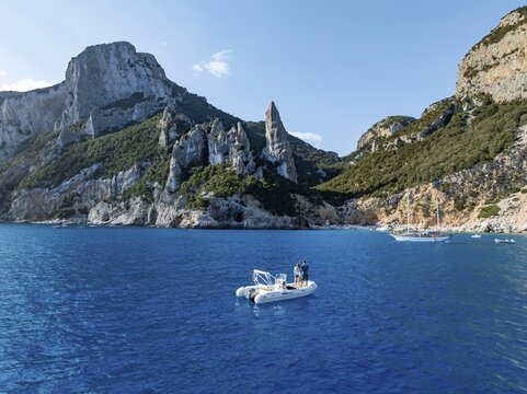 Couple on motorboat off picturesque rocky coast, cliffs with L'Aguglia pinnacle, blue sea and Cala Goloritz&eacute; beach, aerial view, Golfo di Orosei, Baunei, Sardinia, Italy