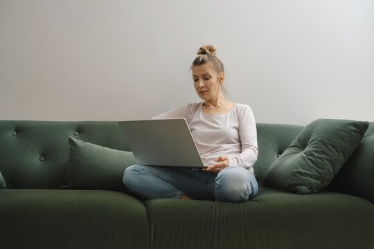 A woman sits cross-legged on a soft green couch, focused on her laptop. Natural light fills the relaxing living room, creating an inviting atmosphere for work or leisure in the afternoon
