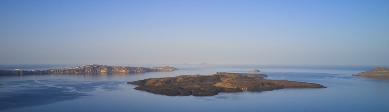 Drone shot, morning atmosphere, caldera, view of volcanic islands Nea Kameni, Palea Kameni and Aspronisi, view of southern tip of the island, sea smooth as glass and blue, sky blue and cloudless, caldera in front of Firostefani, Santorini Island, Cyclades, Greece