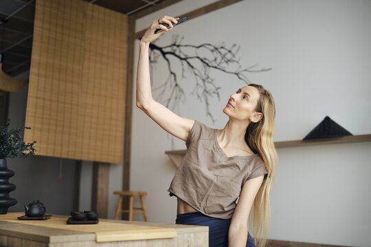 A woman with long hair sits at a wooden table in a simple room. She holds a phone up to take a selfie. The background has some decor and a tree branch print on the wall