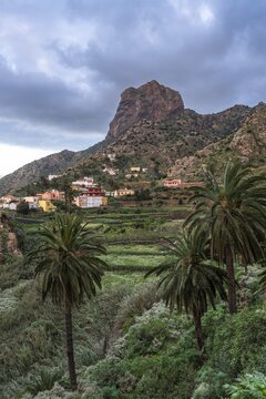 View of Roque El Cano and some houses, palm trees and landscape. Evening, cloudy sky. Vallehermoso, La Gomera, Canary Islands, Spain