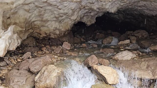 A rocky stream flows over rocky terraces inside a dark snow cave, with sunlight filtering through gaps in the icy ceiling above.
📍 Hawraman Takht, Kurdistan, Iran 