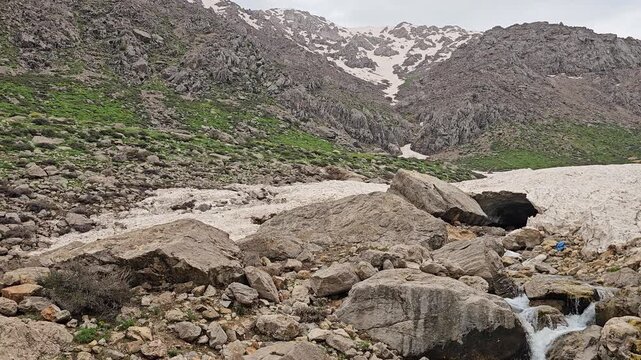A rocky mountain stream flows out from beneath a large avalanche snow patch beside a boulder, with a small snow cave opening, framed by green hillsides and snow-capped rugged peaks.
📍 Hawraman Takht