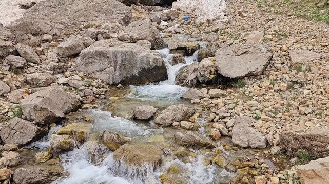 A rocky mountain stream flows out from beneath a large avalanche snow patch beside a boulder, with a small snow cave opening, framed by green hillsides and snow-capped rugged peaks.
📍 Hawraman Takht