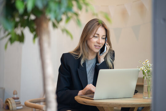 Busy businesswoman with laptop talking on smartphone in coffeeshop coffeehouse with business project. Happy lady solves work issues takes calls. Female employee working remotely