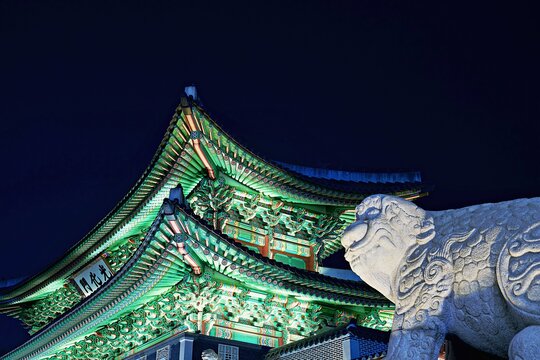A close-up shot of a traditional Korean stone guardian statue (Haetae) flanking the beautifully illuminated colorful eave of Gwanghwamun Gate at night.
