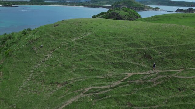 Woman Walking On Green Hilltop, Ocean Backdrop Lone Figure Moves Across Windswept Grass Toward Rocky Promontory, Soft Breeze, Overcast Sky, Wide Aerial Perspective Capturing Scale,Bukit Merese, Lombok
