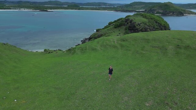 Woman Walking On Green Hilltop, Ocean Backdrop Lone Figure Moves Across Windswept Grass Toward Rocky Promontory, Soft Breeze, Overcast Sky, Wide Aerial Perspective Capturing Scale,Bukit Merese, Lombok