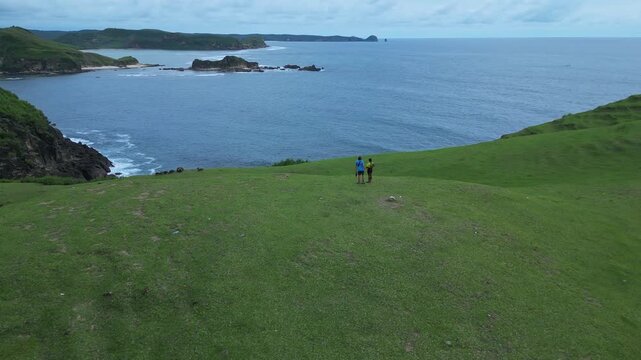 Couple Hiking Along Coastal Ridge Aerial Drone View Showing Two Hikers In Blue And Yellow Jackets Crossing Green Grassy Hill Above Ocean And Rocky Islets, Overcast Sky, Peaceful Travel Mood,Lombok