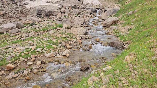 A rocky mountain stream flows through a valley beneath patches of avalanche snow, framed by boulders and green hillsides leading toward rugged snow-capped peaks.
📍 Hawraman Takht, Kurdistan, Iran 