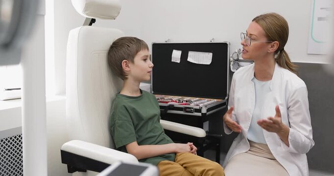 Friendly female children eye doctor in white coat talking to little boy patient after examination explaining test results giving advice on preventing eyesight problems discussing healthy vision habits