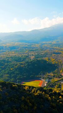 Vertical video. Nicolosi, Sicily, Italy. Mount Etna, volcanic craters overgrown with dense forest, aerial panoramic view under a bright blue sky with scattered clouds. Drone footage. Rich colors