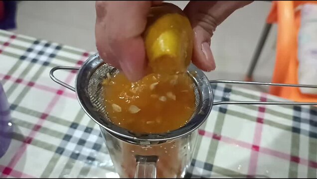 Hand squeezing yellow lemon through a metal strainer into a glass on a checkered tablecloth, making fresh homemade citrus juice.