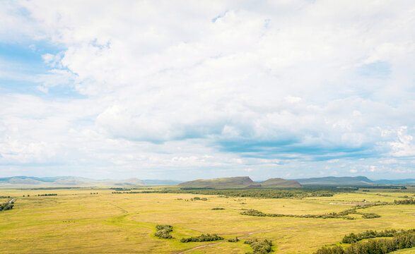Aerial photography from drone, steppe valley with yellow-green meadows, dirt road, groves of bushes, gently rolling mountains on horizon, cloudy sky