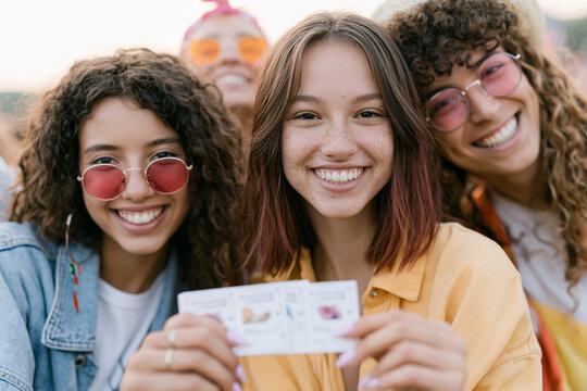Three smiling young women pose together outdoors. They wear colorful sunglasses and casual clothes. One holds a small booklet or card with pictures. Their joyful expressions convey friendship and fun