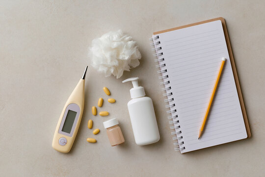 Minimalist setup for health tracking and self-care routines. Digital thermometer rests beside pills and lotion. Notebook, pencil, and fluffy bath puff suggest wellness habits