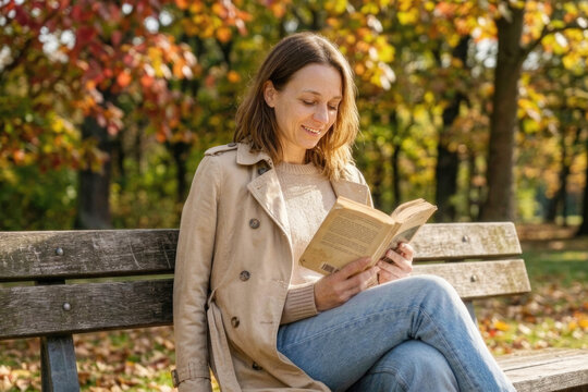 Happy woman in trench coat reading book on wooden park bench during autumn