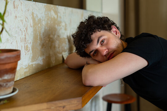 A young man rests his head on his crossed arms while leaning against a wooden counter. He looks directly at the camera in an indoor setting.