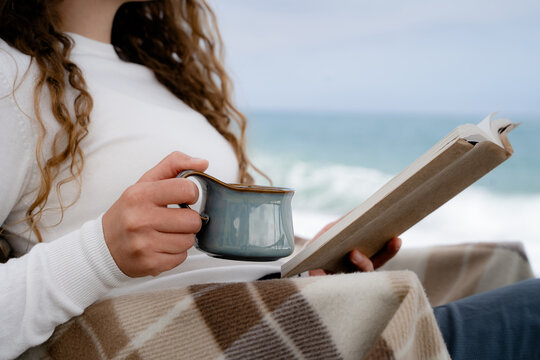 Close-up of a female hand holding a warm drink and open book, while relaxing by the ocean. Mindfulness, comfort, and peaceful outdoor recreation.