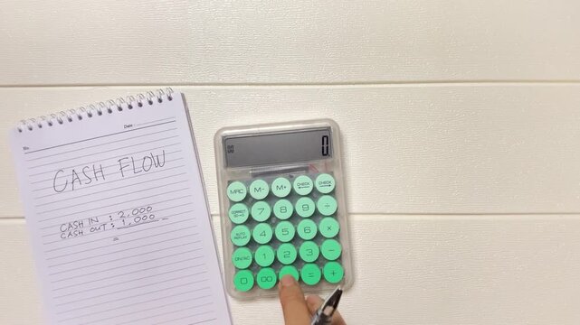 Top view of a male business owner calculating cash flow using a calculator while writing financial records in a notebook with a pen. Wearing eyeglasses, this organized workspace represents business