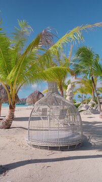 Vertical Screen: Bora Bora tropical beach resort featuring white wicker cage lounge pod on white sand surrounded by palm trees and thatched huts with turquoise lagoon water in background.